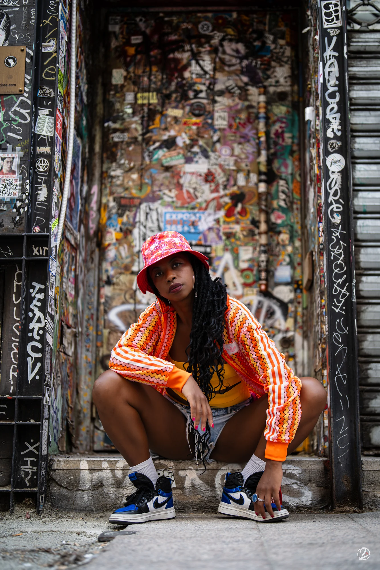 Portrait devant une porte couverte de graffitis, veste orange et bob coloré.
