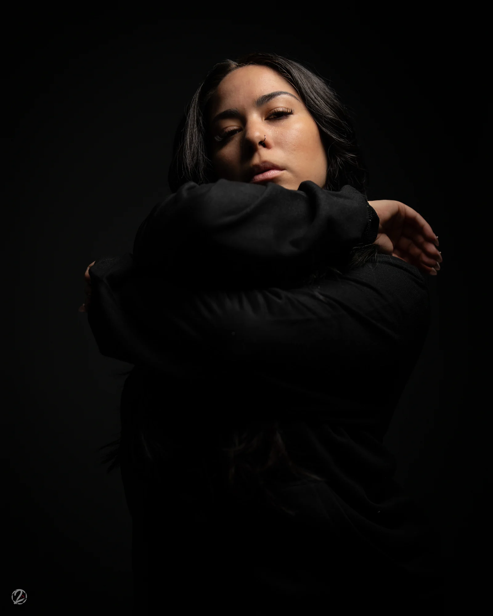 Portrait en tenue noire, bras relevé sur fond sombre.